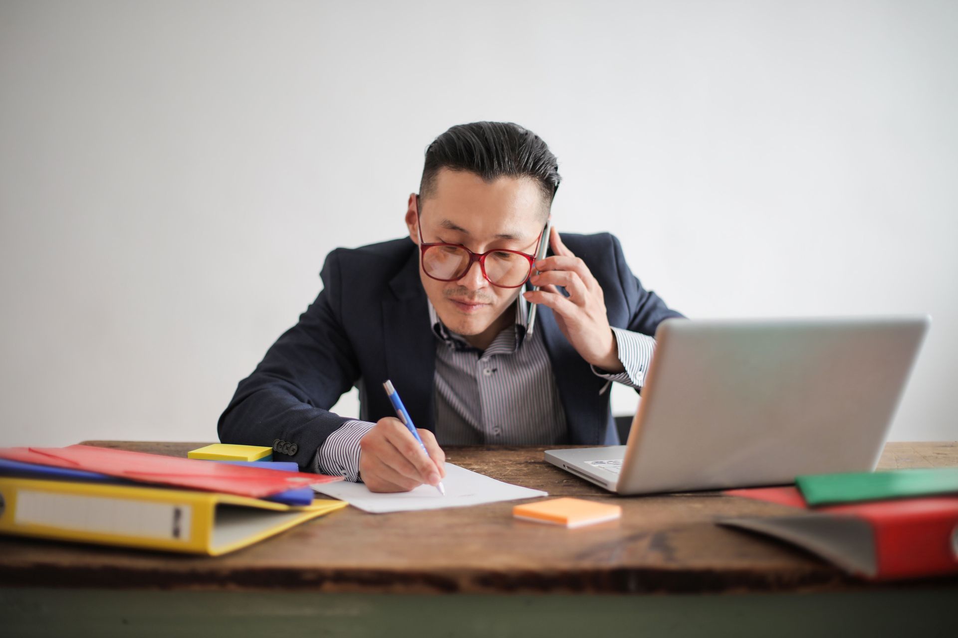 Man writing at a desk with papers, laptop, and books, while talking on a phone