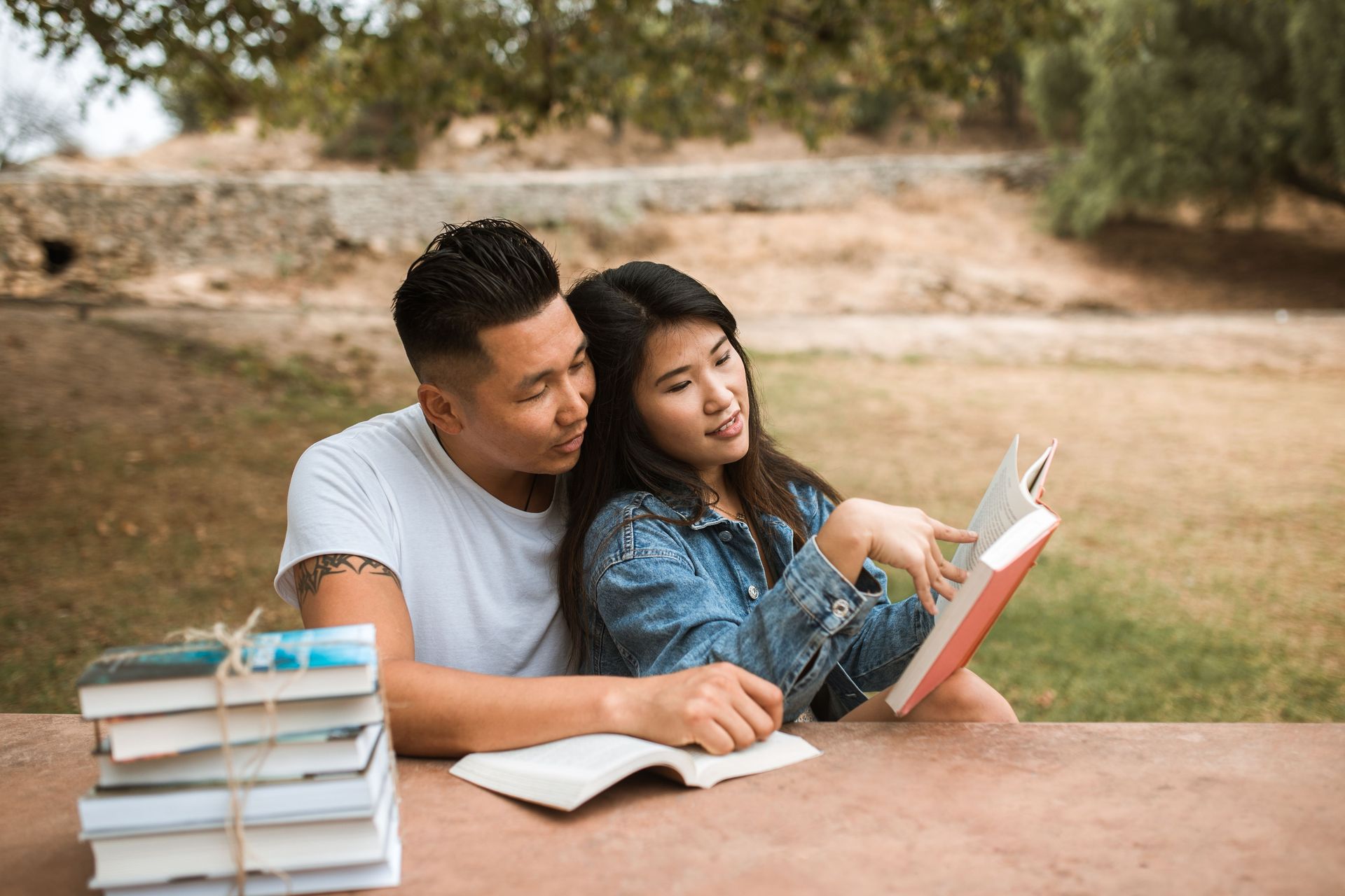 Two people reading a book outdoors at a table with a stack of books nearby