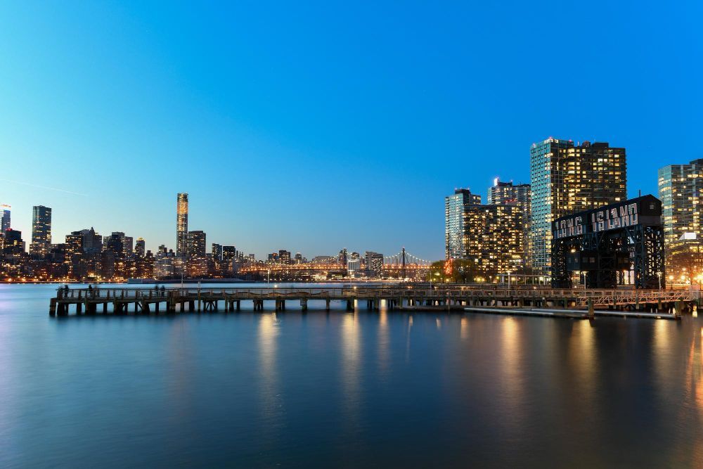 City skyline at dusk reflected on calm water, with lit high-rises and a pier in the foreground
