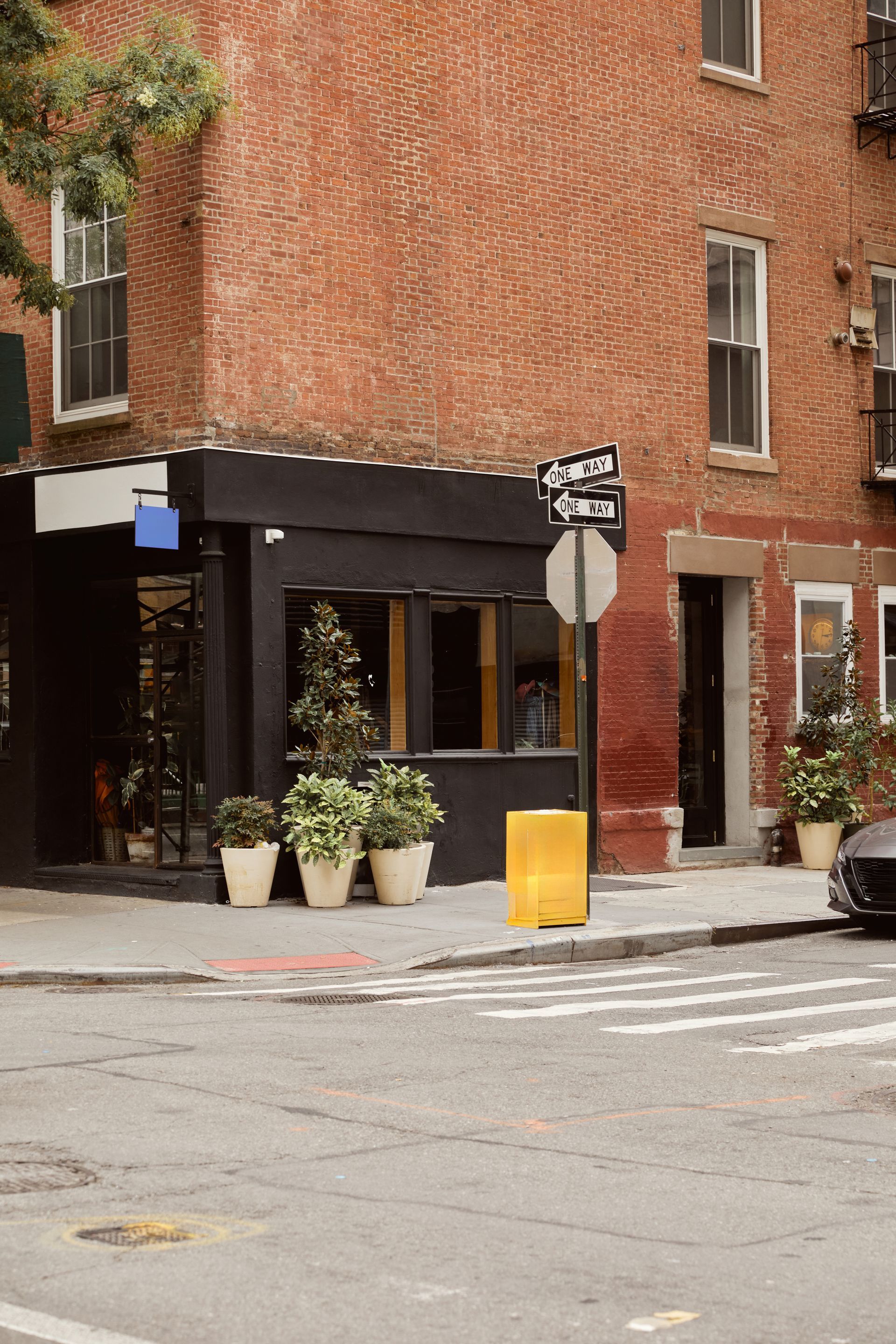 Brick street corner with a black awning, potted plants, and a yellow traffic bollard beside a crosswalk