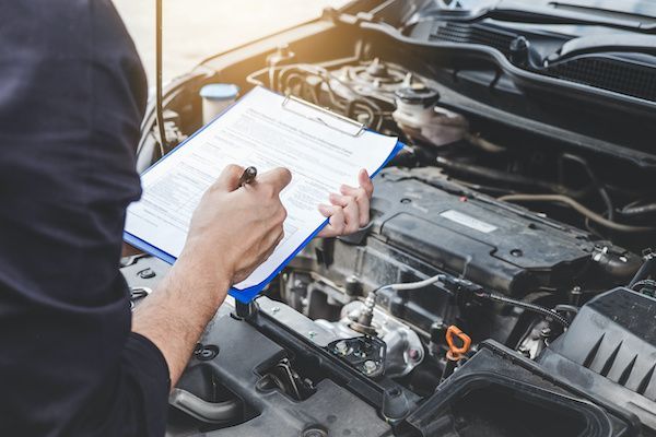 A man is writing on a clipboard while looking under the hood of a car. | Lex Brodies 