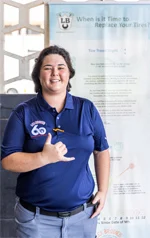 A woman in a blue shirt is giving a thumbs up while standing in front of a sign. | Lex Brodies
