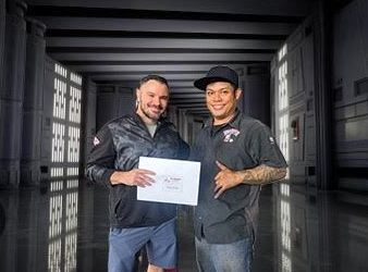 Two men are standing next to each other in a hallway holding a piece of paper. | Lex Brodies