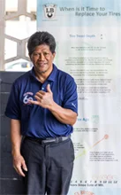 A man in a blue shirt is standing in front of a sign that says `` when is it time to replace your tires ''. | Lex Brodies