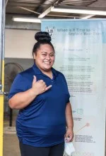 A woman in a blue shirt is standing in front of a sign and giving a thumbs up. | Lex Brodies