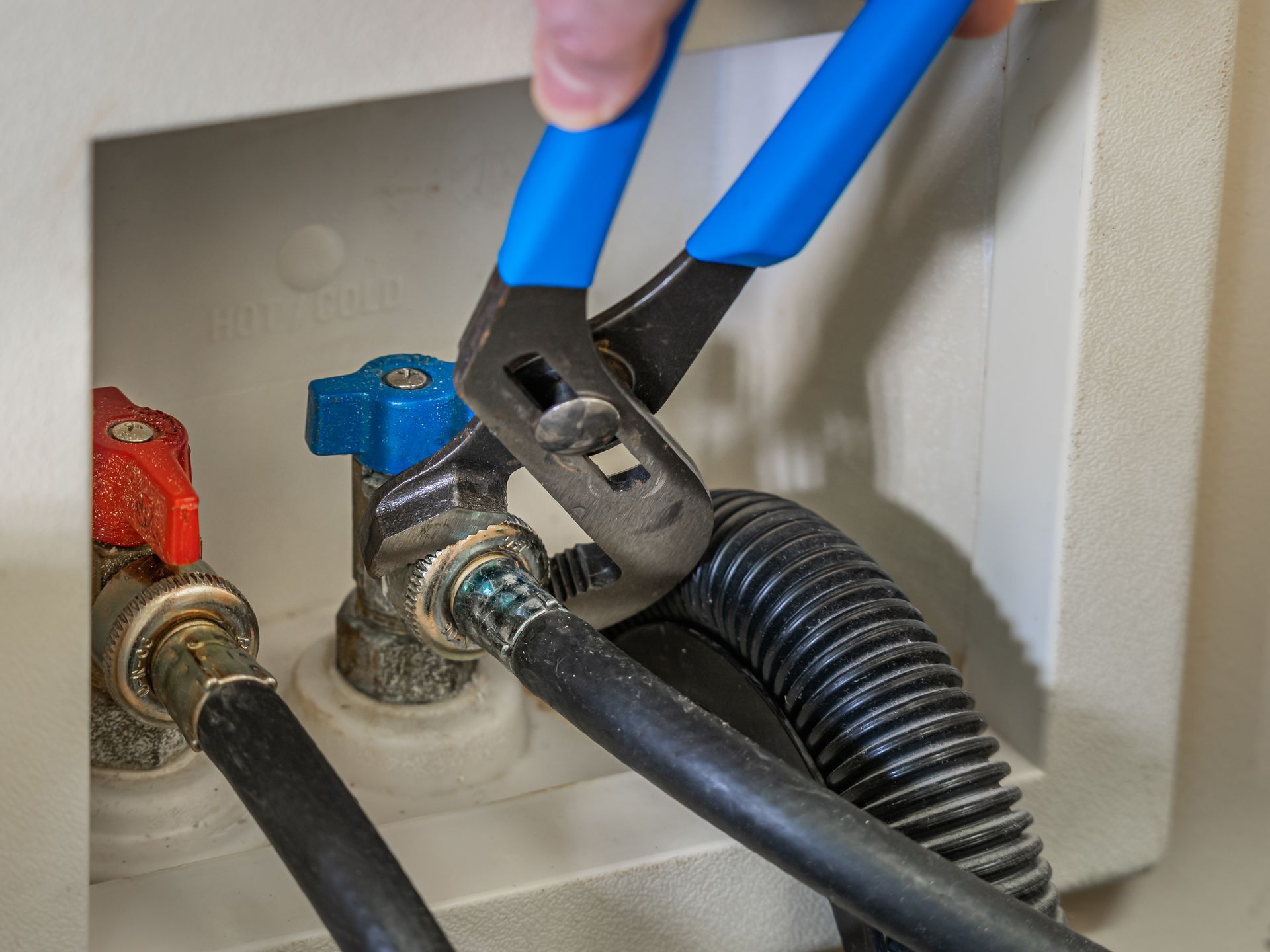 A person uses pliers to loosen a washing machine hose connection to a water valve.