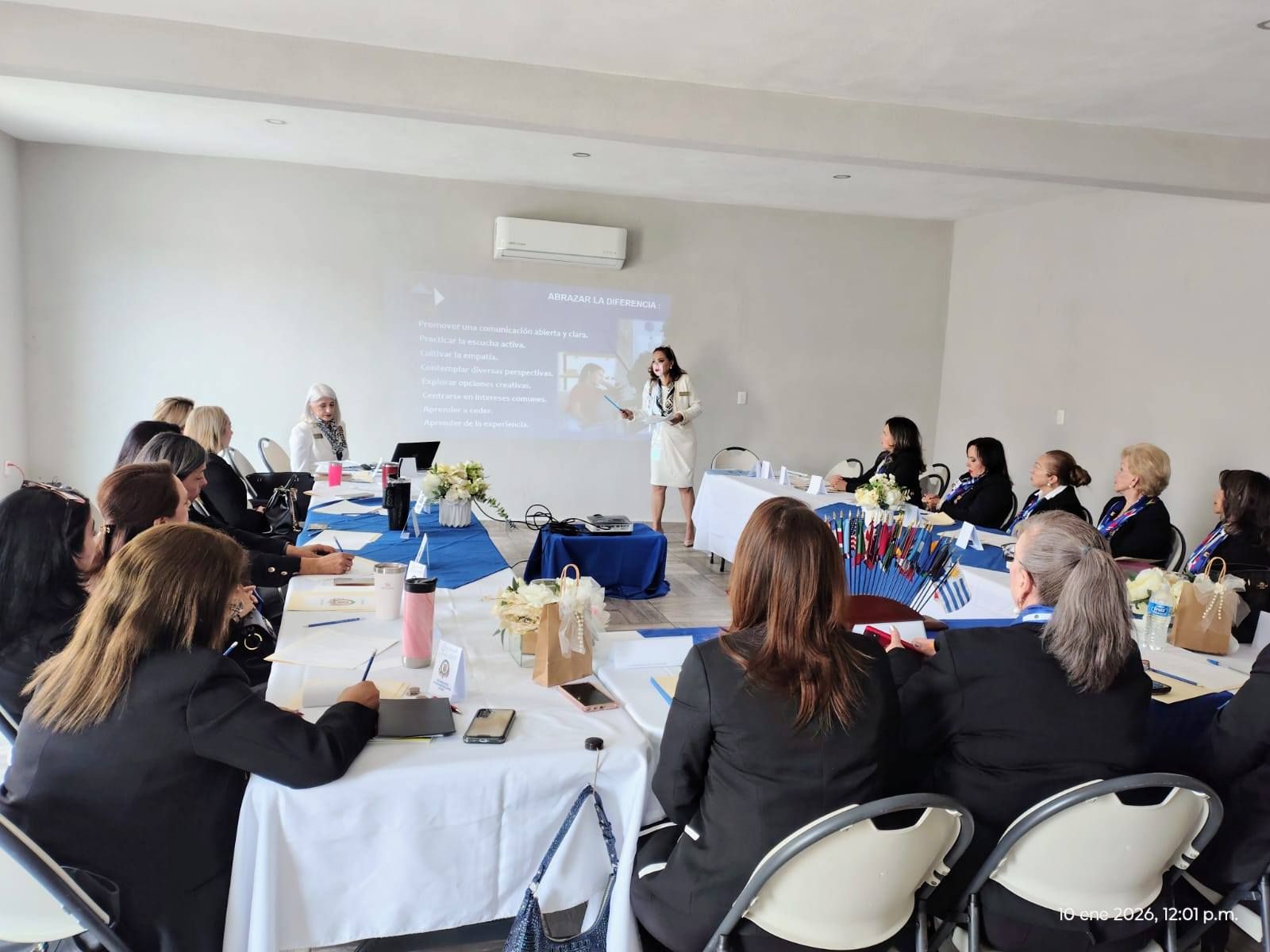 Woman presenting to a group in a brightly lit room. Attendees seated at tables. Projector screen visible.