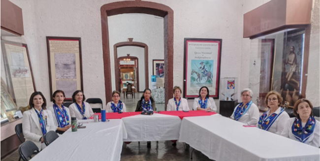 Group of women in white jackets and blue scarves seated at a table in a room with artwork.