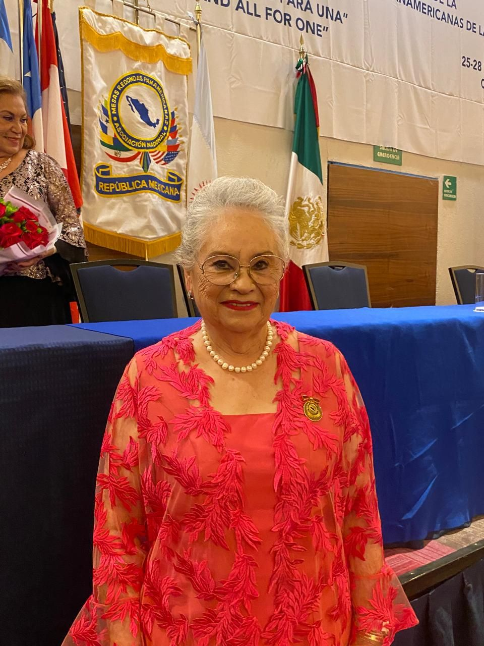 Woman in pink lace jacket stands at podium, flags in background.