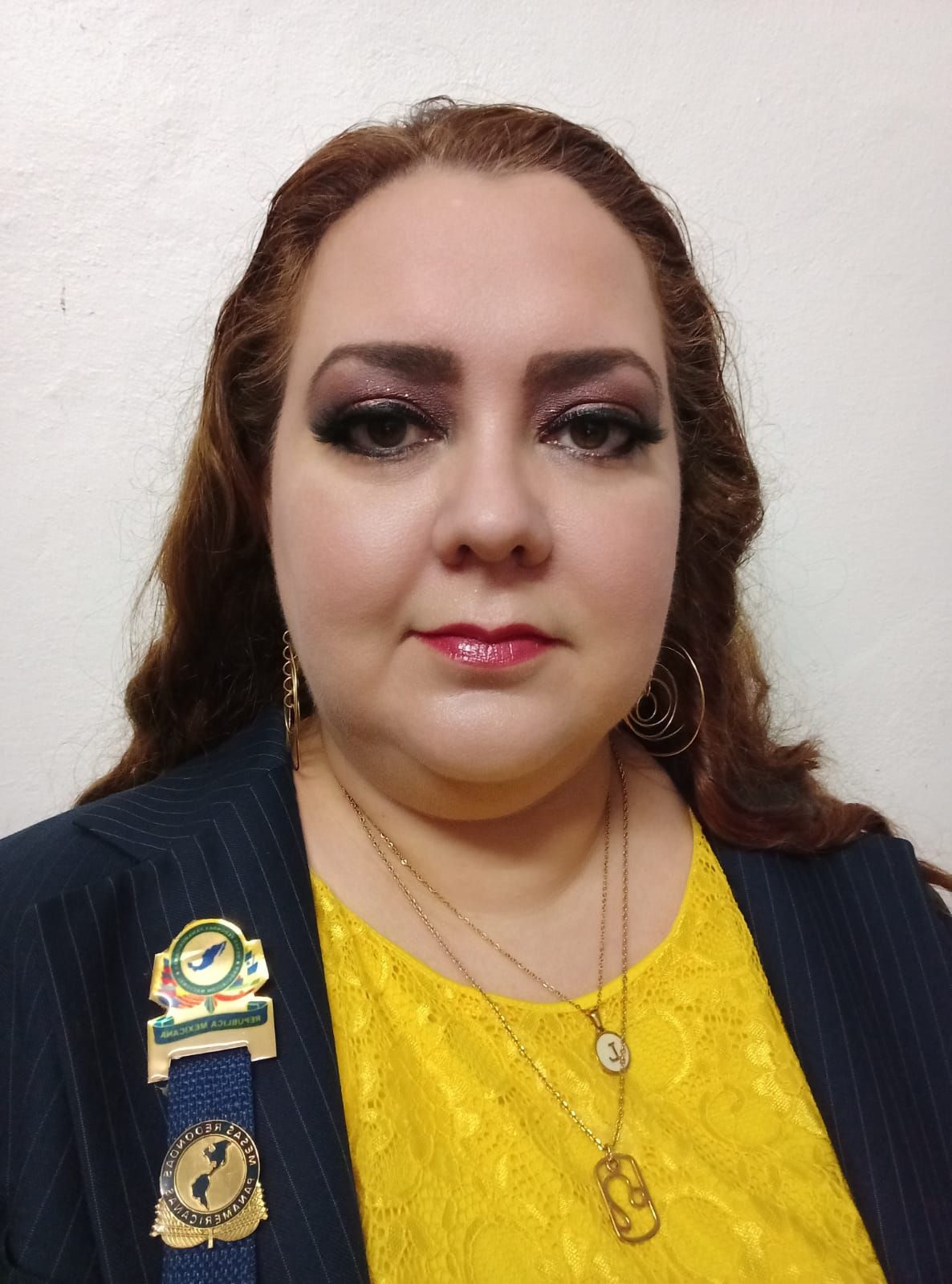 Woman with curly brown hair, wearing a yellow top, blue jacket, and pins.