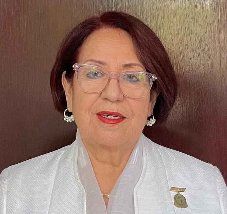 Woman wearing glasses and a white blazer, smiling, in front of a wood background.