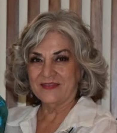 Woman with gray, curly hair smiles, wearing a white shirt; wooden backdrop.