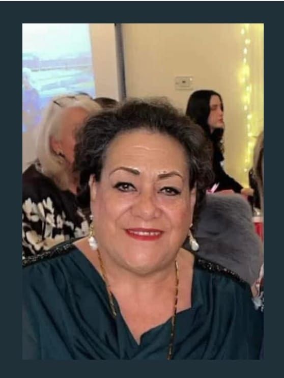 Woman with dark hair smiles, wearing a green top and pearl earrings, in a social setting.
