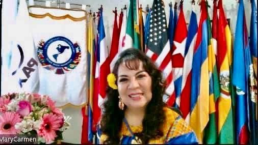 Woman in front of flags, smiles, wears a flower and colorful top. Flowers in front of her.