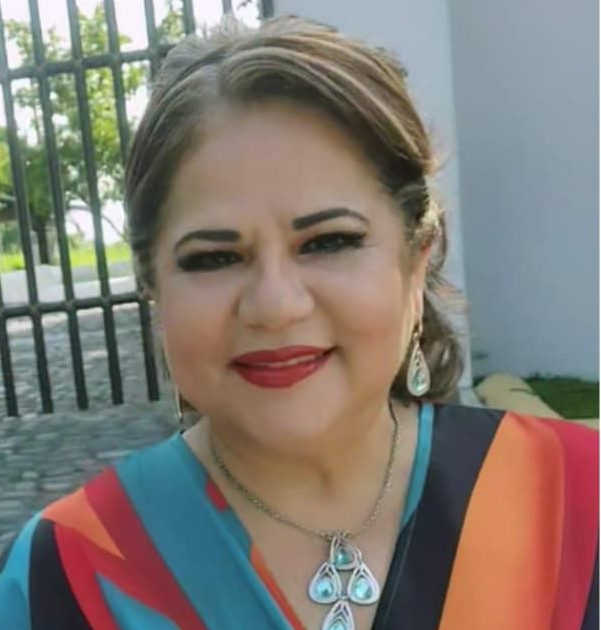 Woman with dark hair, wearing a patterned top and necklace, smiling near a wrought-iron fence.