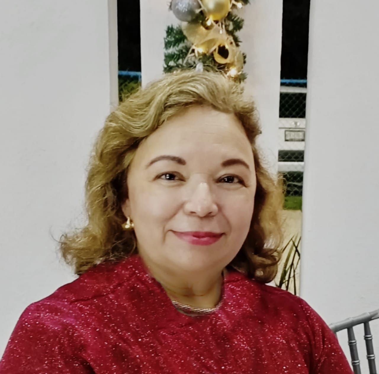 Woman in red top smiles; curly hair, gold earrings, Christmas decorations behind.