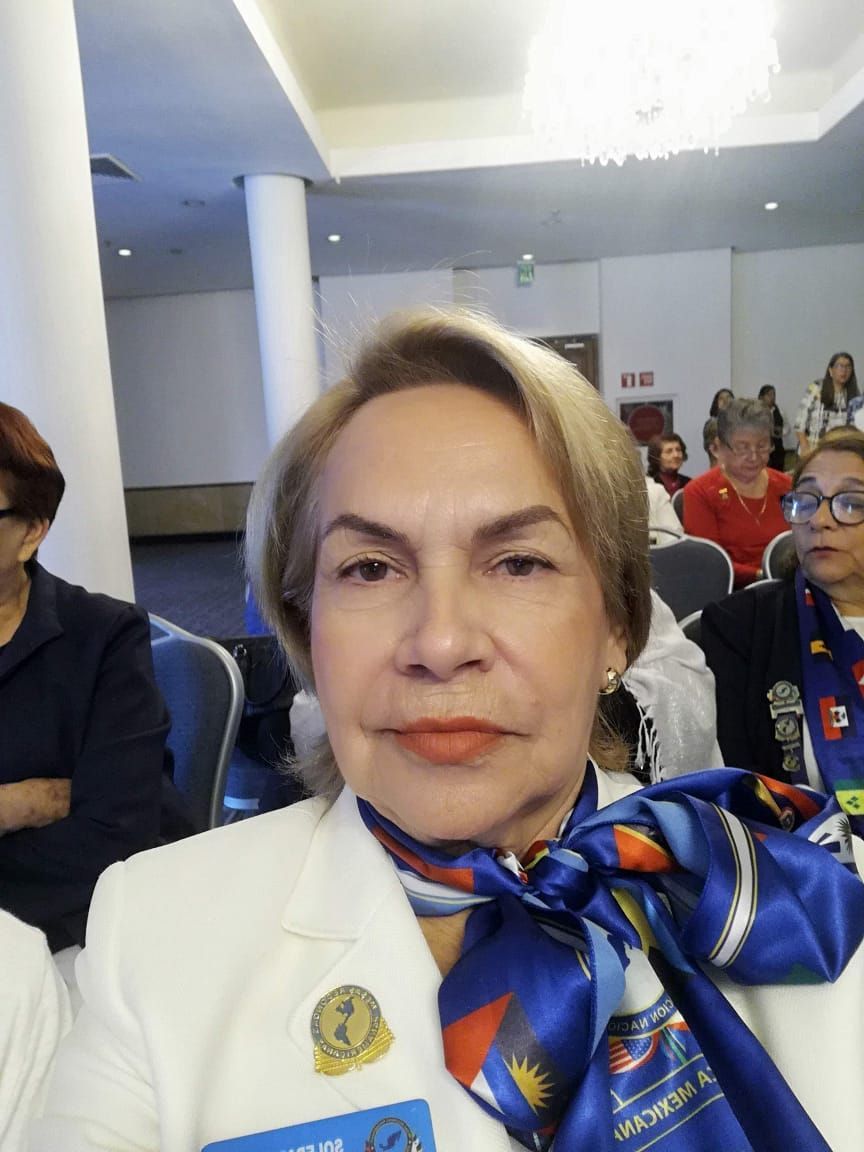 Woman in white suit, blue scarf, and pin, seated at a conference or event.