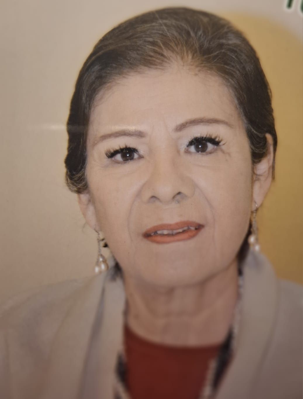 Woman with dark hair and makeup, wearing a red top and gray jacket, looking at the camera.