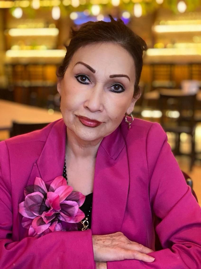 Woman in pink blazer smiles, wearing flower brooch. Indoors setting, neutral background.