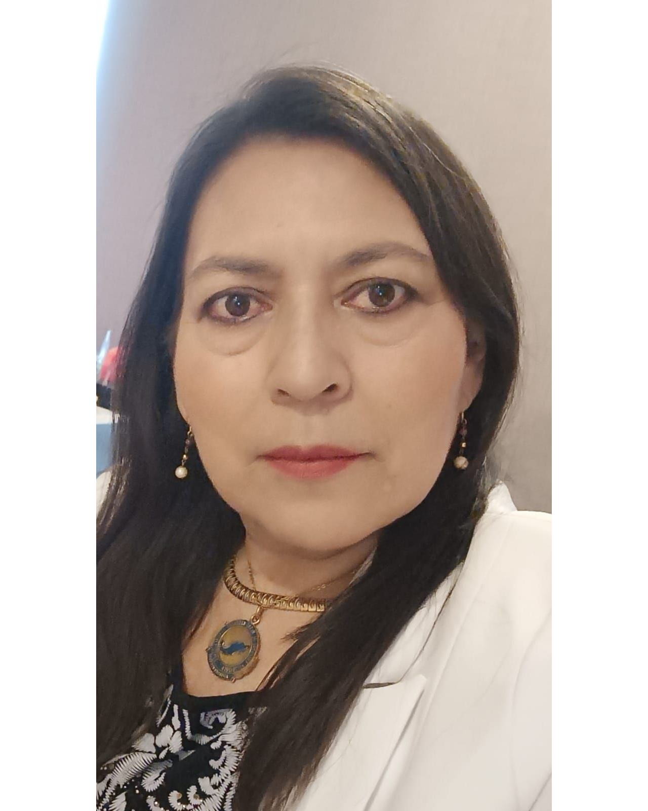 Woman with long dark hair wearing a white blazer, gold necklace, and earrings, looking at the camera.