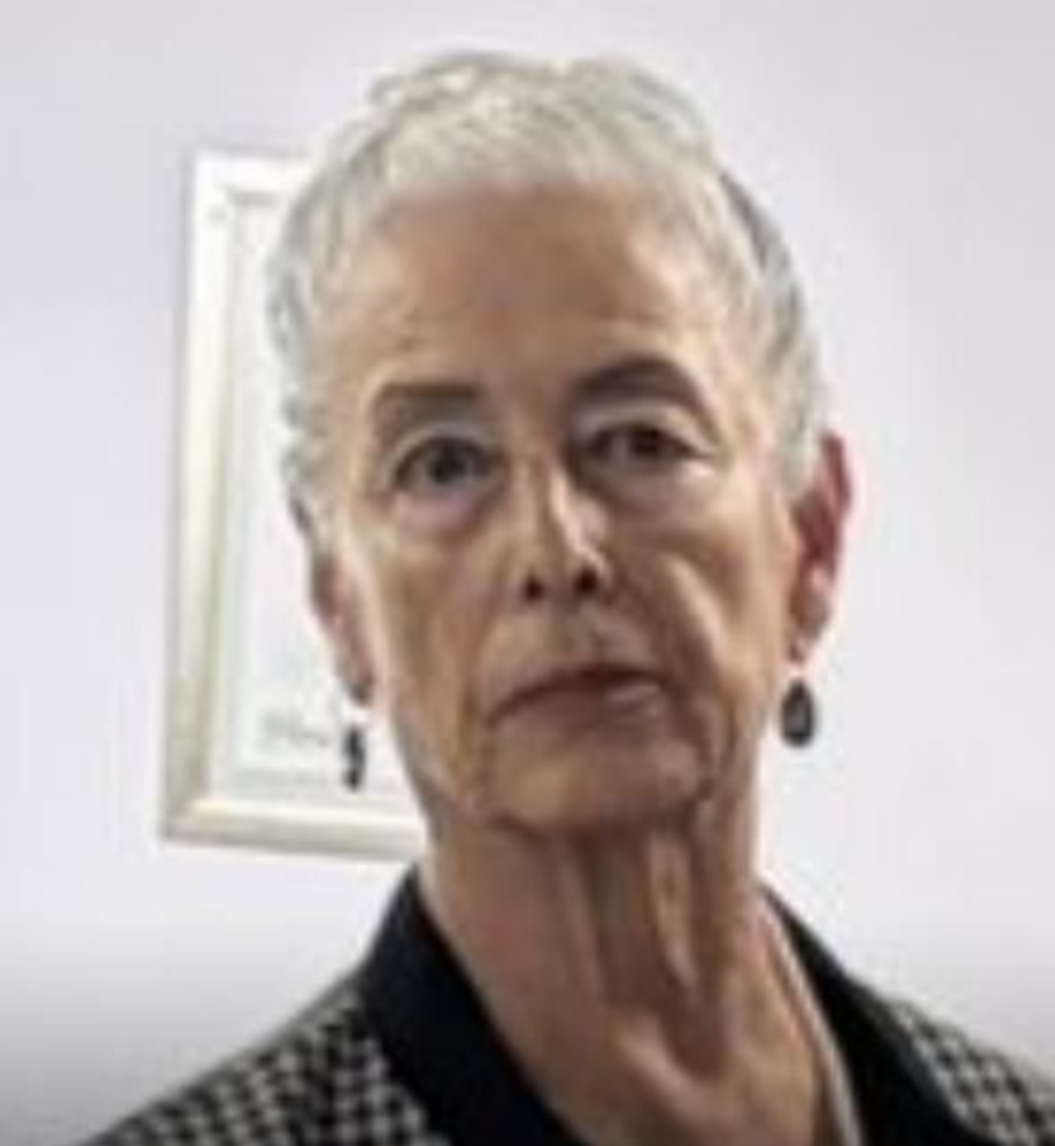 Woman with short gray hair, wearing a black and white patterned jacket and earrings, stands in front of a framed picture.