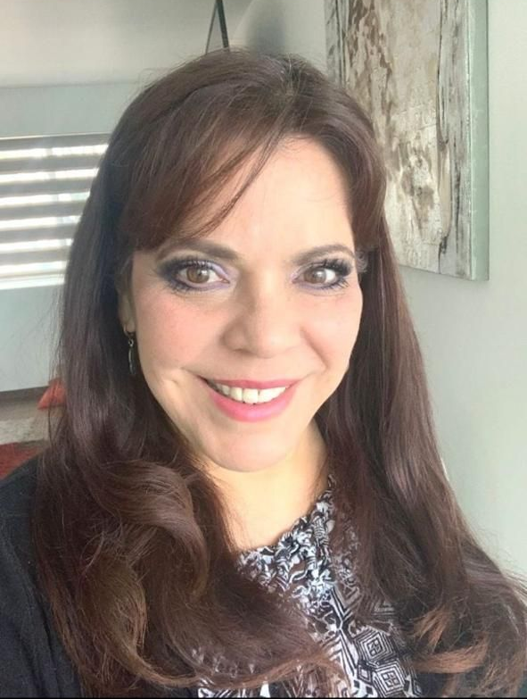 Woman with long brown hair, smiling, wearing a patterned top, in front of a window with blinds.