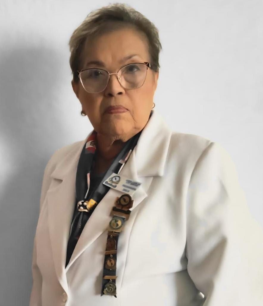 Woman wearing glasses and a white blazer, looking at the camera. She has pins on her lapel and a scarf around her neck.