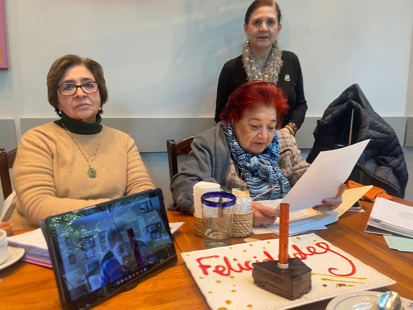 Three women at a table. One reads a paper, another looks forward, and the third stands behind them. A cake and tablet are on the table.