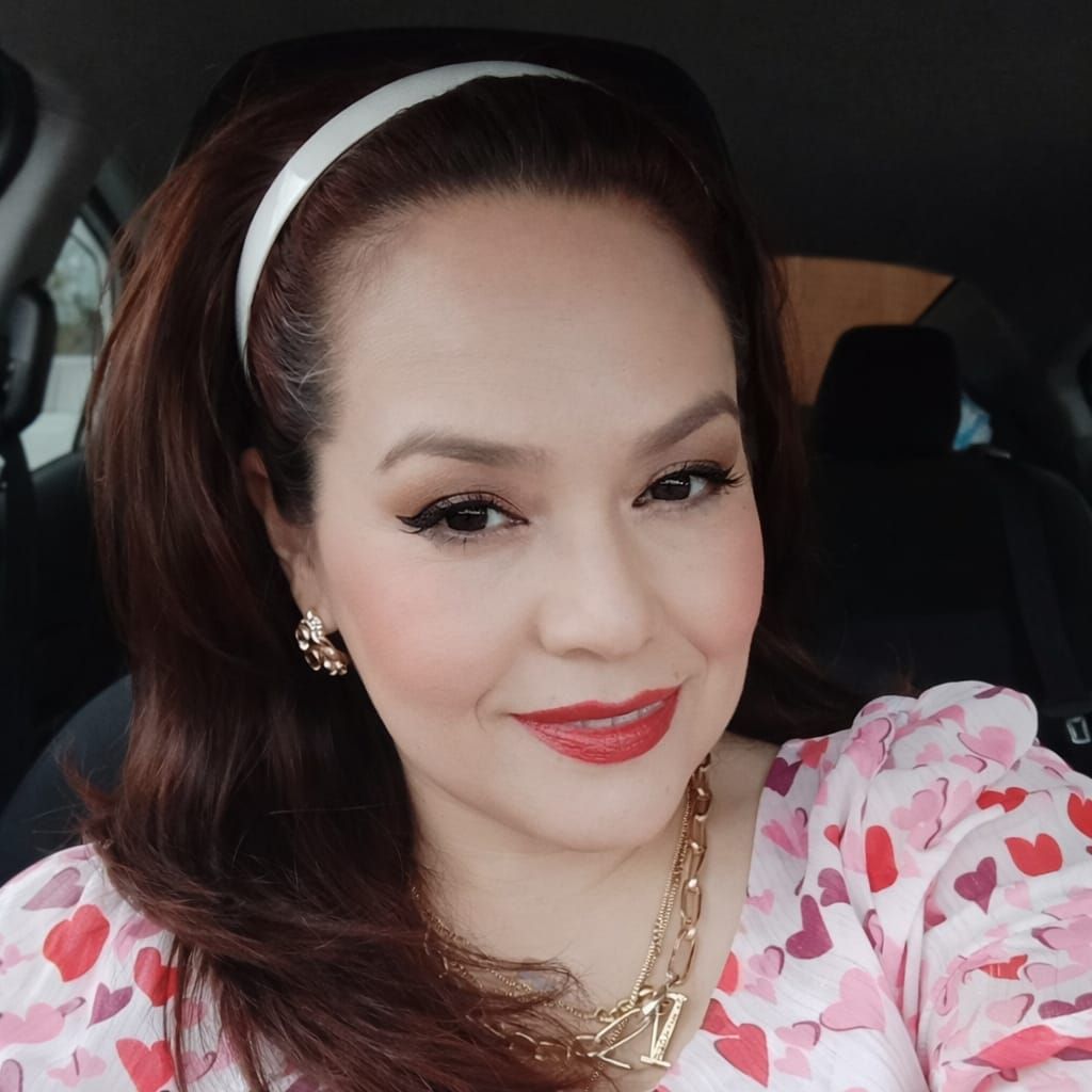 Woman in a car wearing a white headband, red lipstick, and a pink floral top smiles.