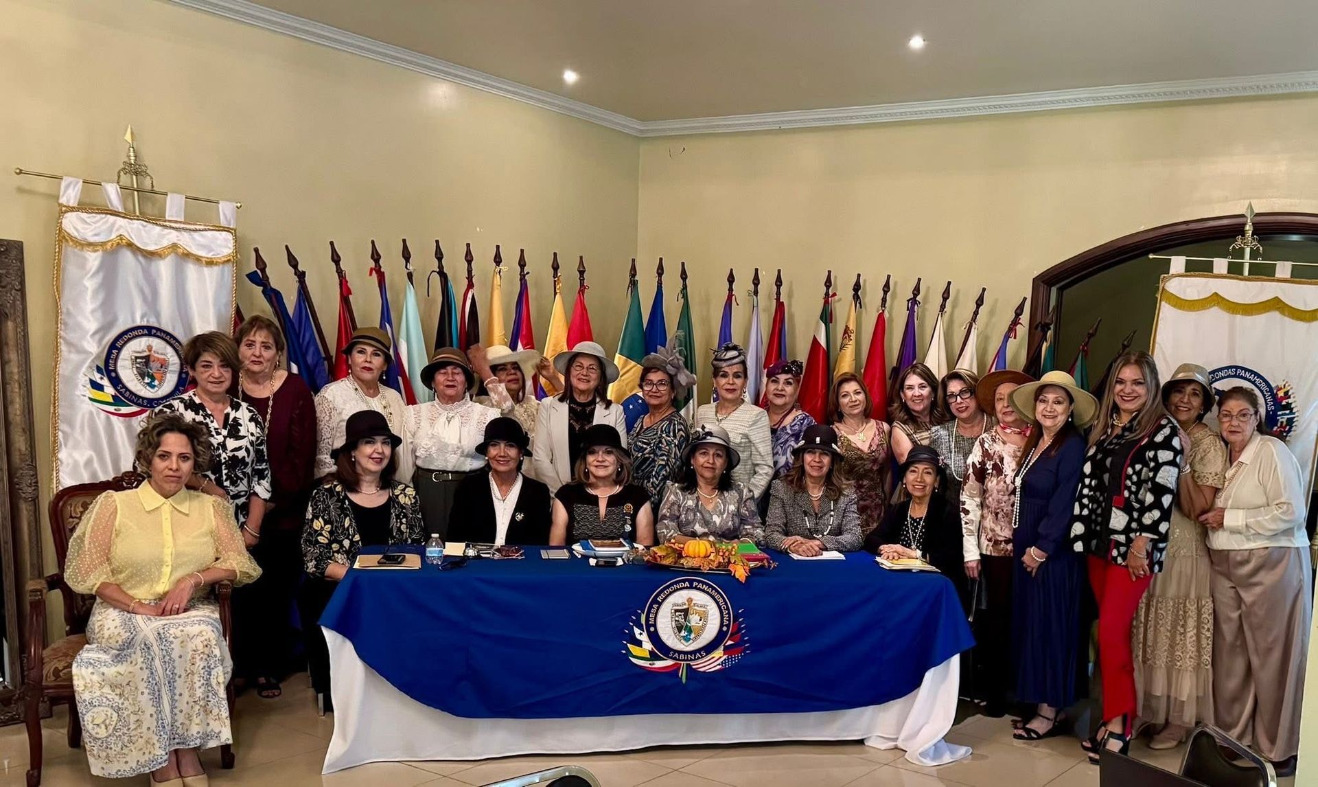 Group of women gathered behind a table with a blue tablecloth, flags in the background, formal attire.