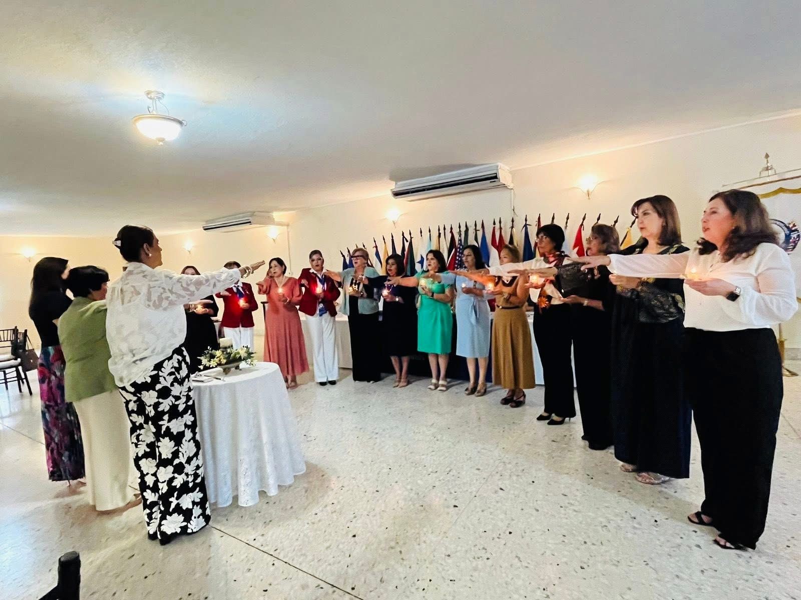 Group of people holding candles, indoors near flags. Women in formal attire are circled around a table.