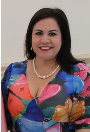 Woman smiling, wearing a colorful floral print dress and a pearl necklace.
