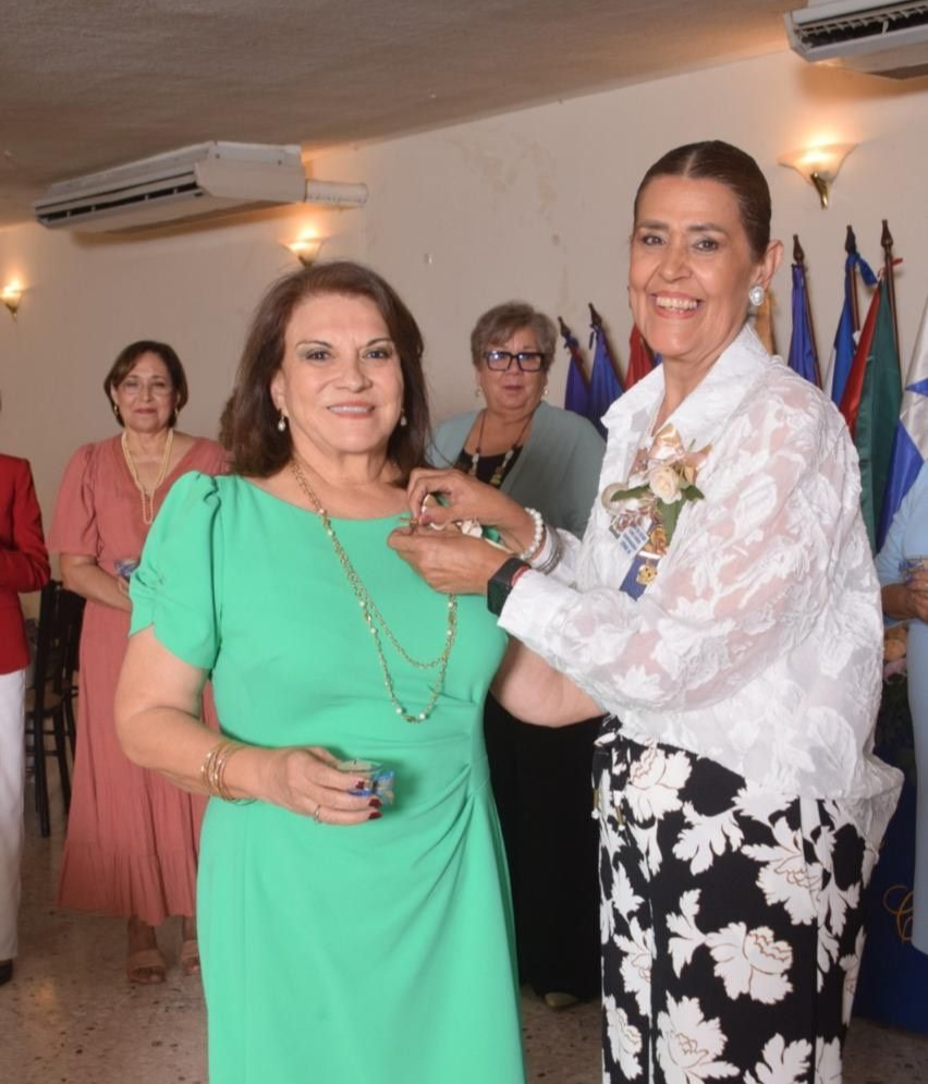 Woman in green dress receives a medal from woman in white jacket; room setting with others present.