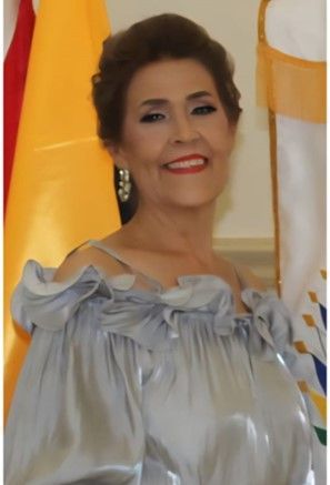 Woman in a silver off-the-shoulder dress smiles. She stands near flags indoors.