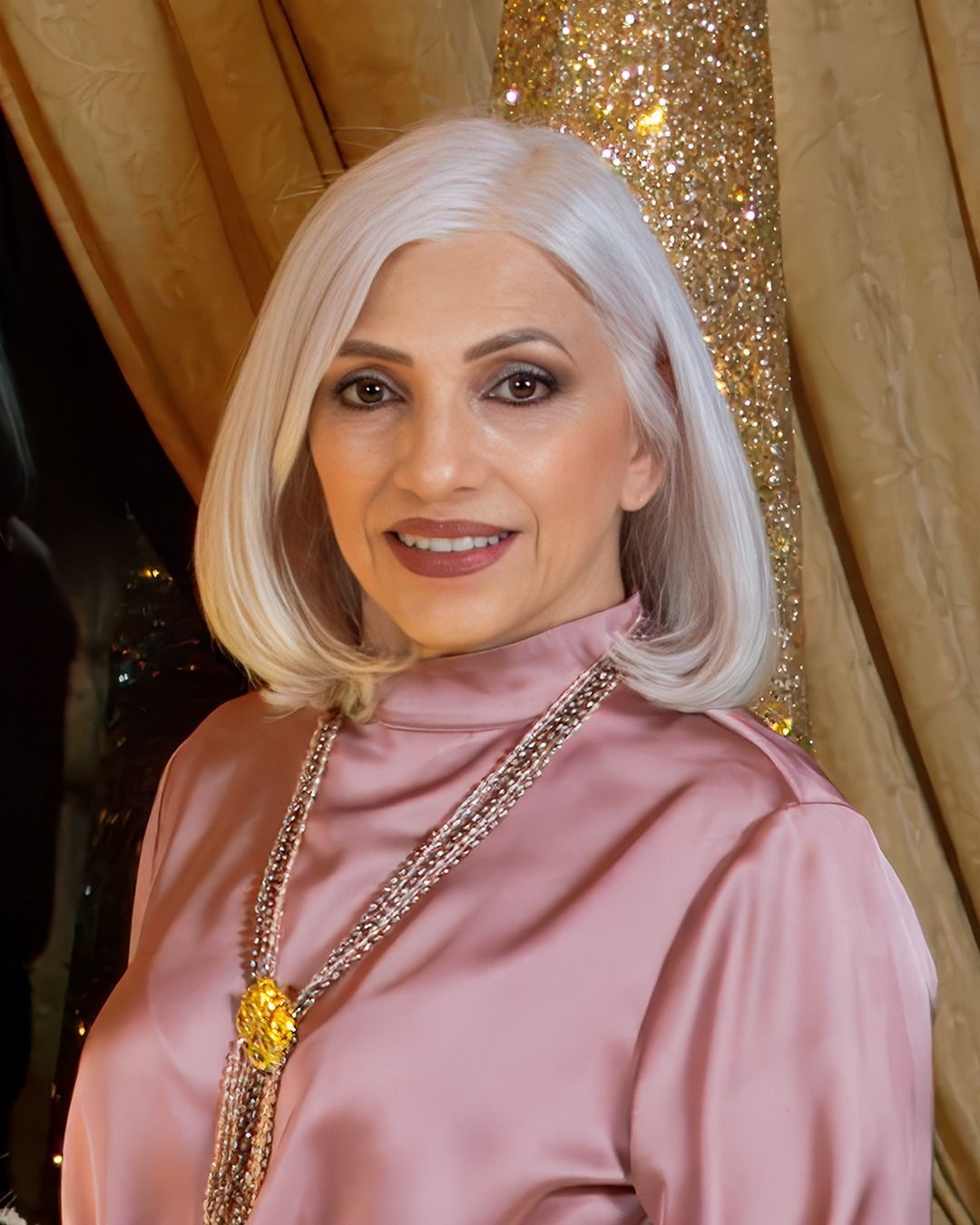 Woman with gray hair smiles, wearing a pink silk top and jeweled necklace, posing indoors.