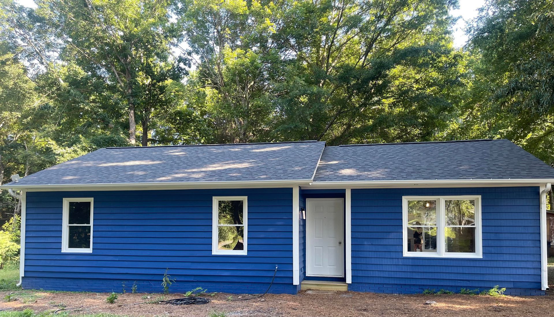 A blue house with white windows and a black roof is surrounded by trees.