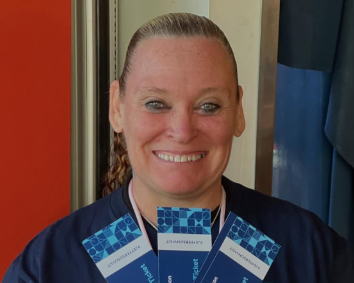 Person in navy shirt and visor smiles, holding heart-shaped pepperoni pizza in a shop.