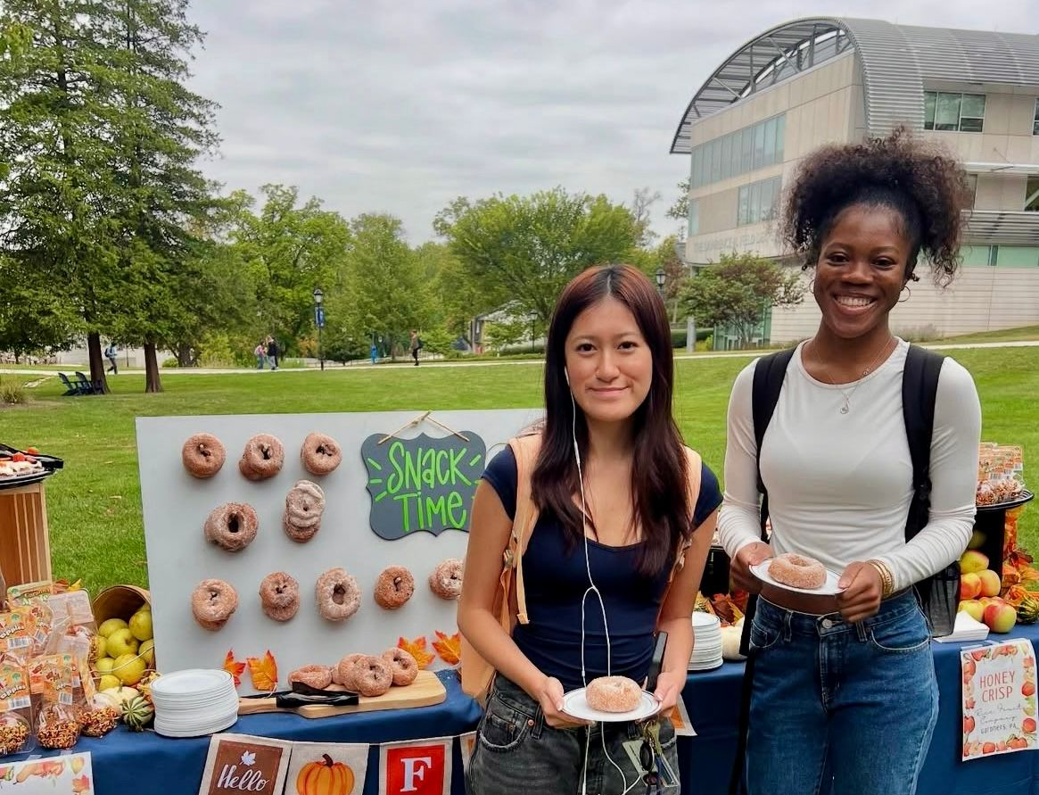 Two students with donuts at an outdoor snack table, fall decorations, campus background.
