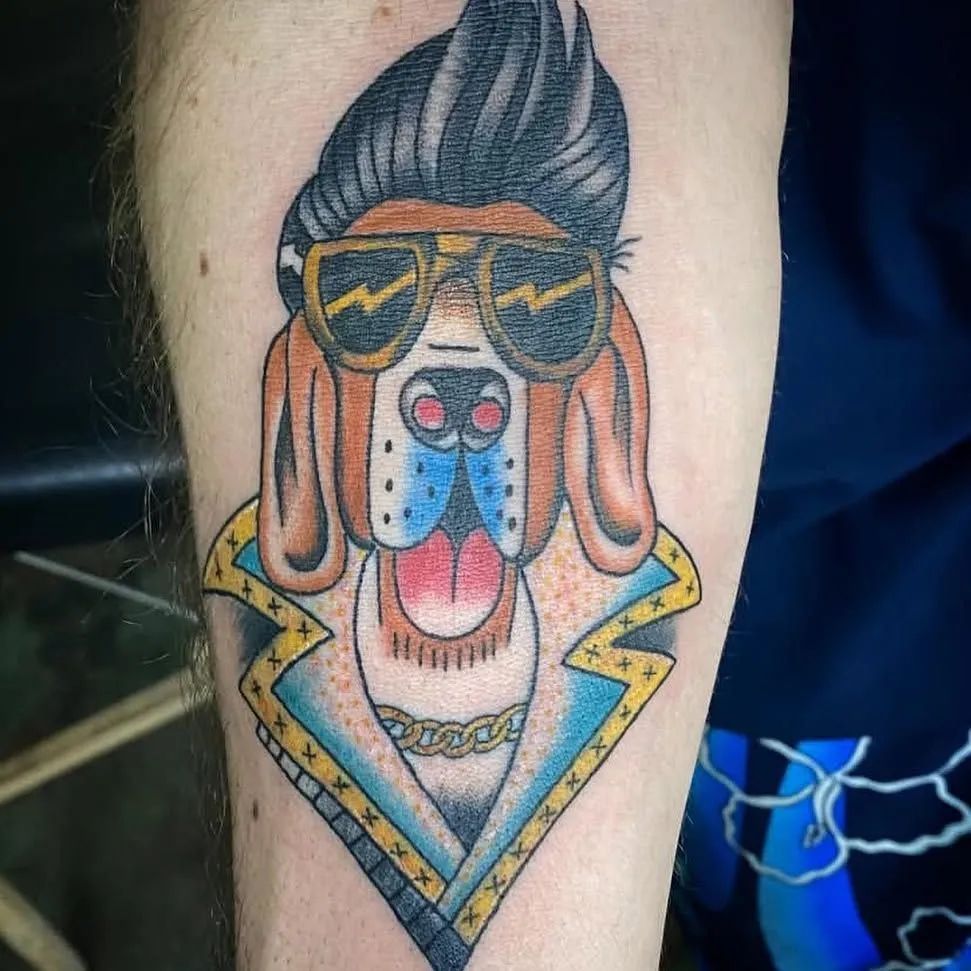 A tattoo of a dog with a pompadour hairstyle, sunglasses, and a colorful jacket, wearing a gold chain.