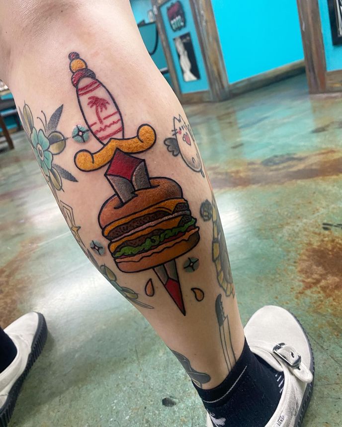 A person has a tattoo of a hamburger and a dagger on their leg.