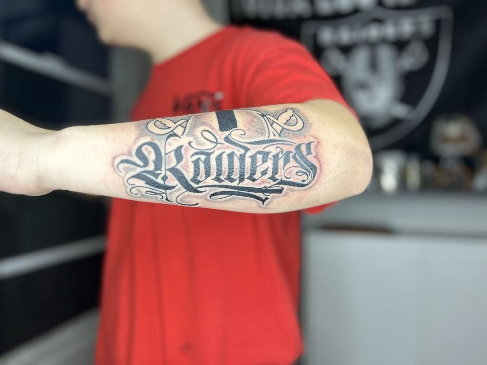 A man in a red shirt has a tattoo of the word raiders on his arm.
