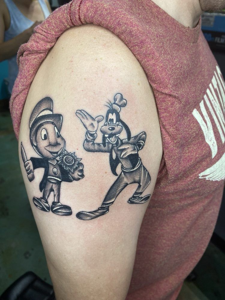 A person has a tattoo of pinocchio and goofy on their arm.