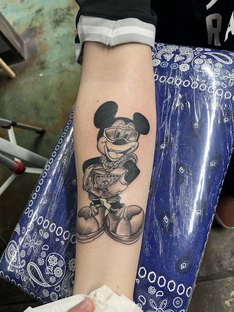 Black and white Mickey Mouse tattoo on forearm, wearing sunglasses, holding money.