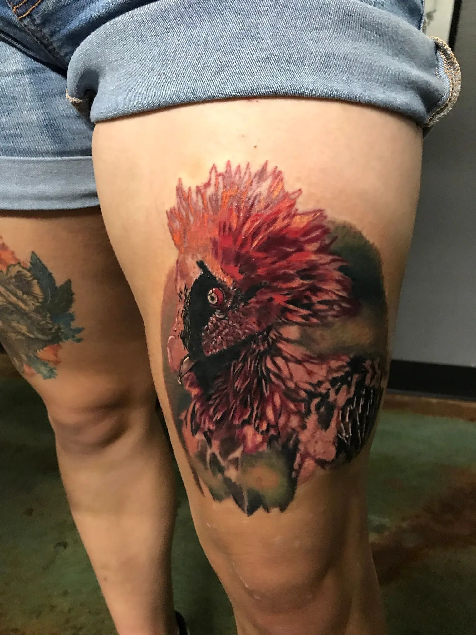 Tattoo of a bird with red plumage on a person's thigh. Dark shading creates depth.