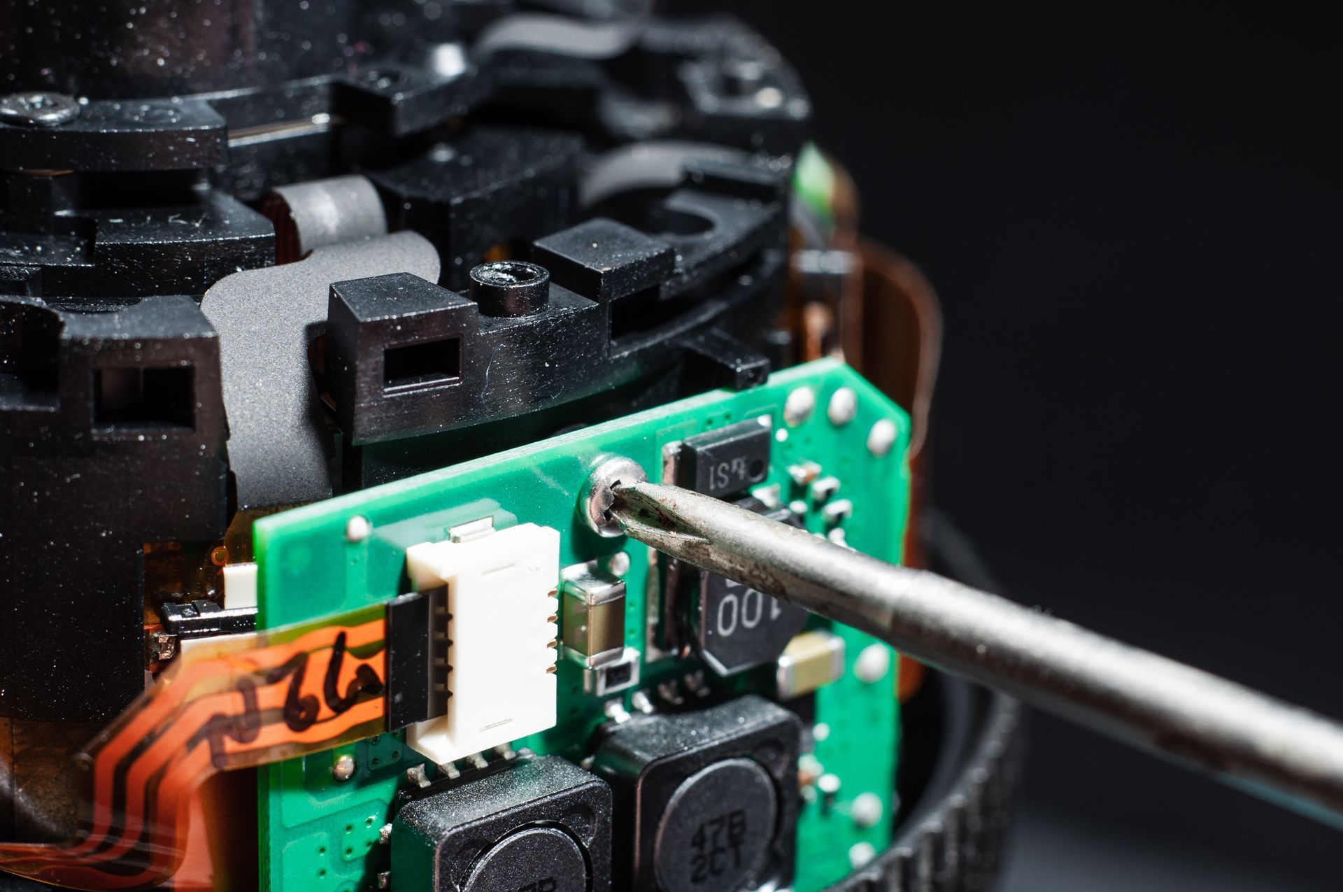 A metal screwdriver bit is positioned over a screw on a green circuit board inside a disassembled electronic device.