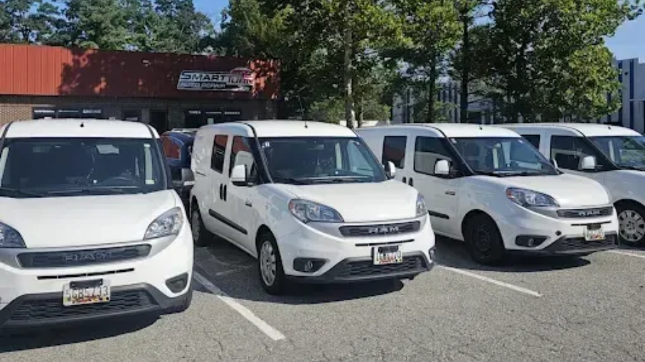 A row of four white RAM ProMaster City vans parked in an outdoor lot in front of a building on a sunny day.