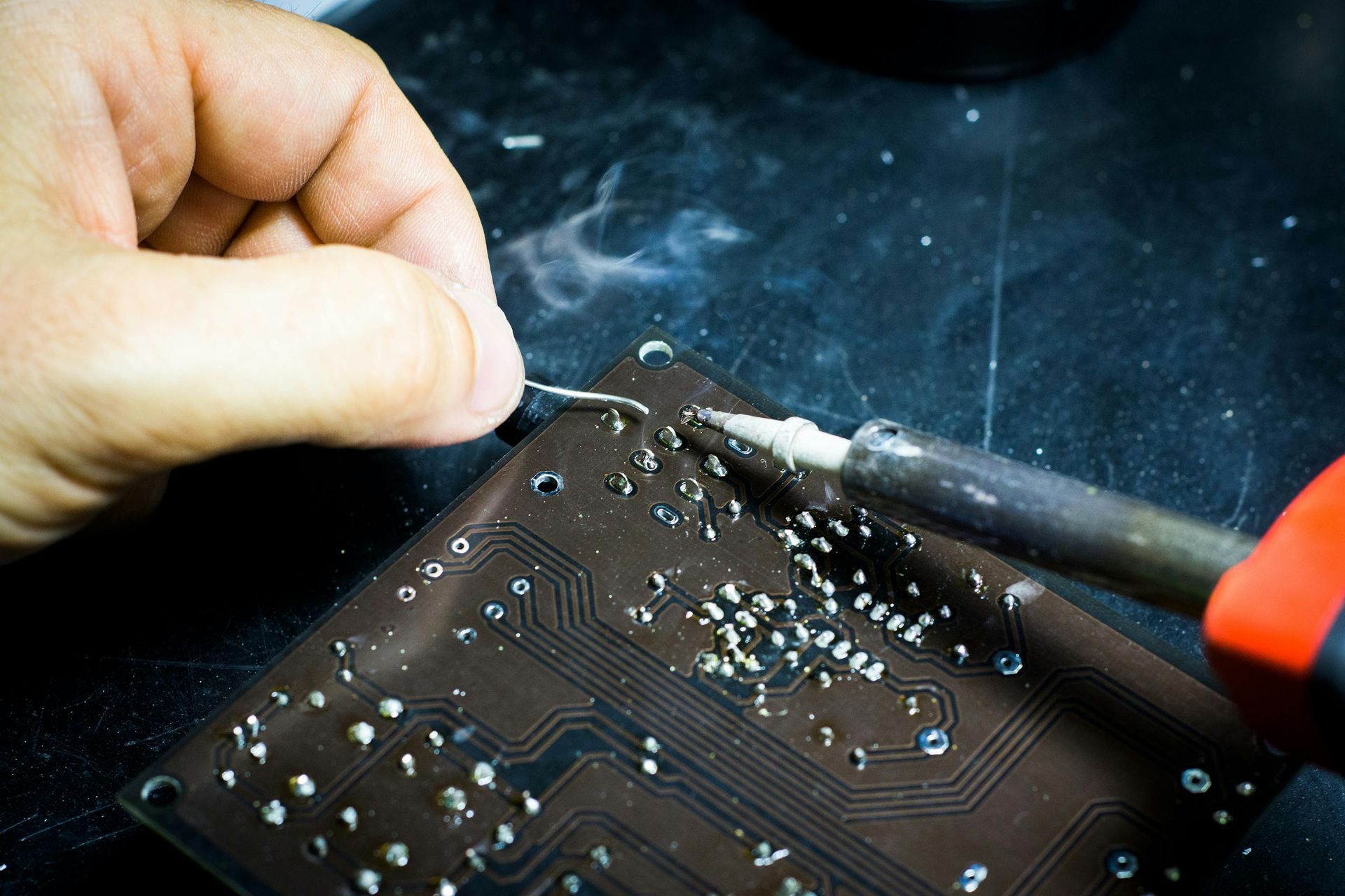 A person uses a soldering iron to attach a component to a circuit board, with visible smoke rising from the connection.