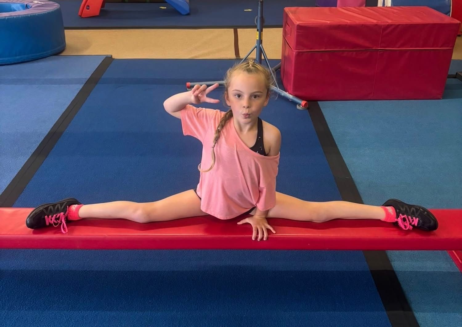 Girl in splits on red beam, pink shirt, giving peace sign, in a gym.