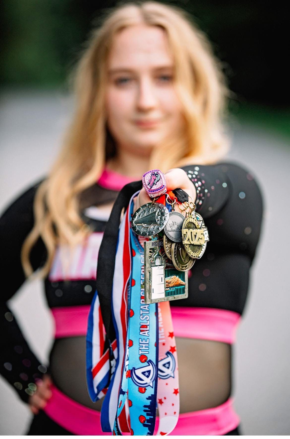 Blond person in cheerleading uniform holds out arm displaying several medals. Pink and black outfit.