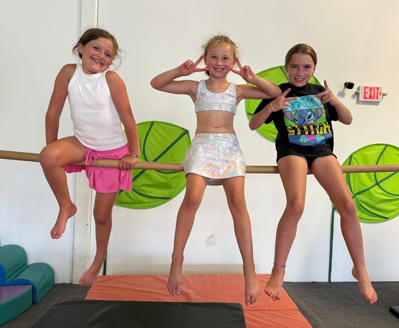 Three girls in activewear on a bar, making playful gestures. Green backdrop.